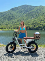 Woman standing next to a small electric bike by a lake with mountains in the background